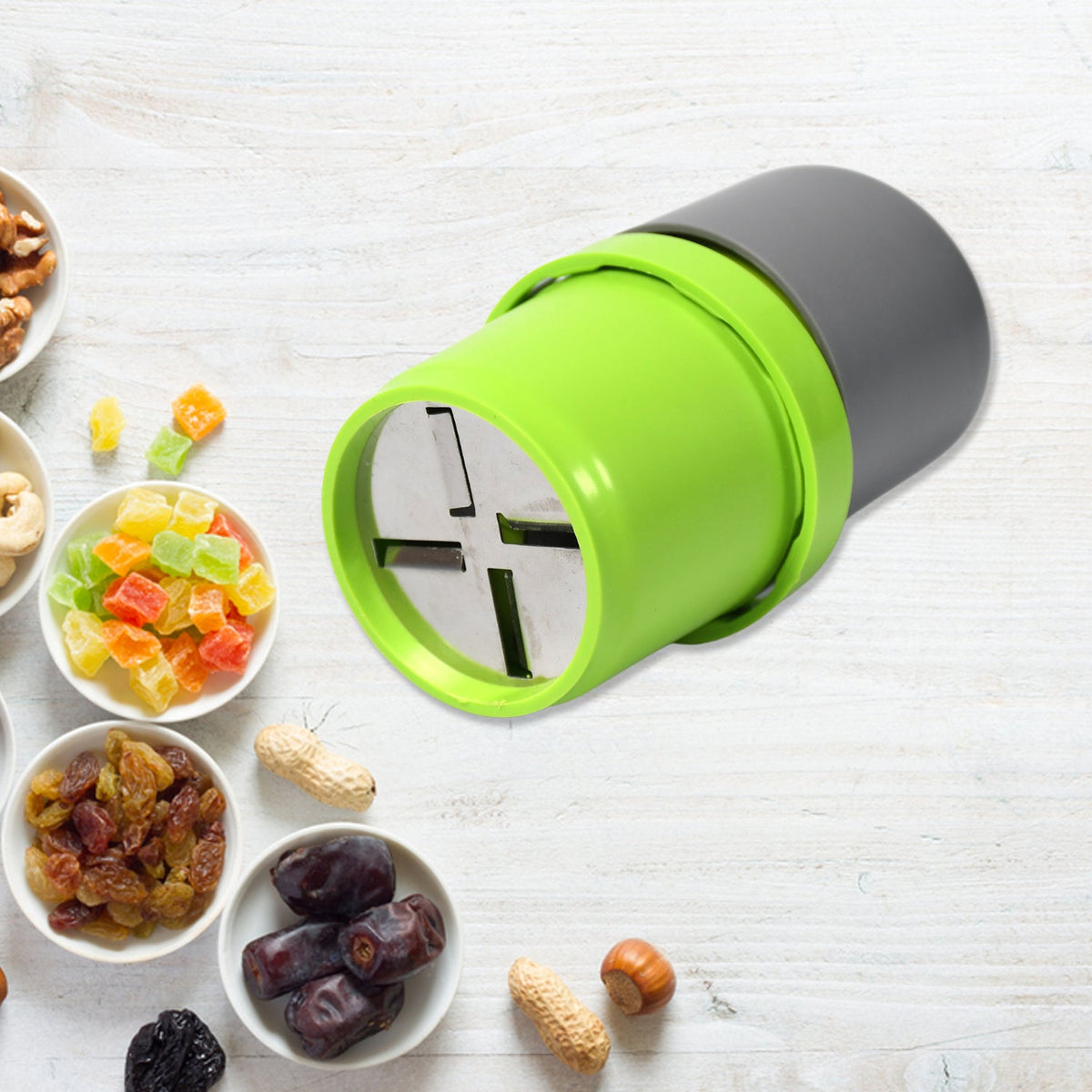 A green and gray dry fruit slicer is centered on a white wooden background, surrounded by small bowls of various dry fruits.
