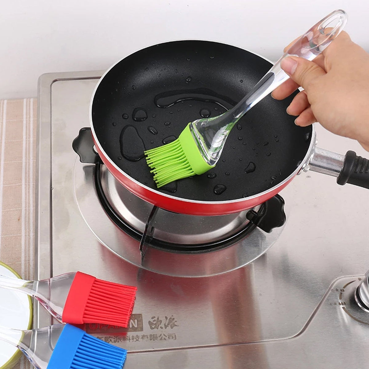 A hand uses a green silicone brush to spread oil in a black frying pan on a gas stove, with red and blue brushes nearby.