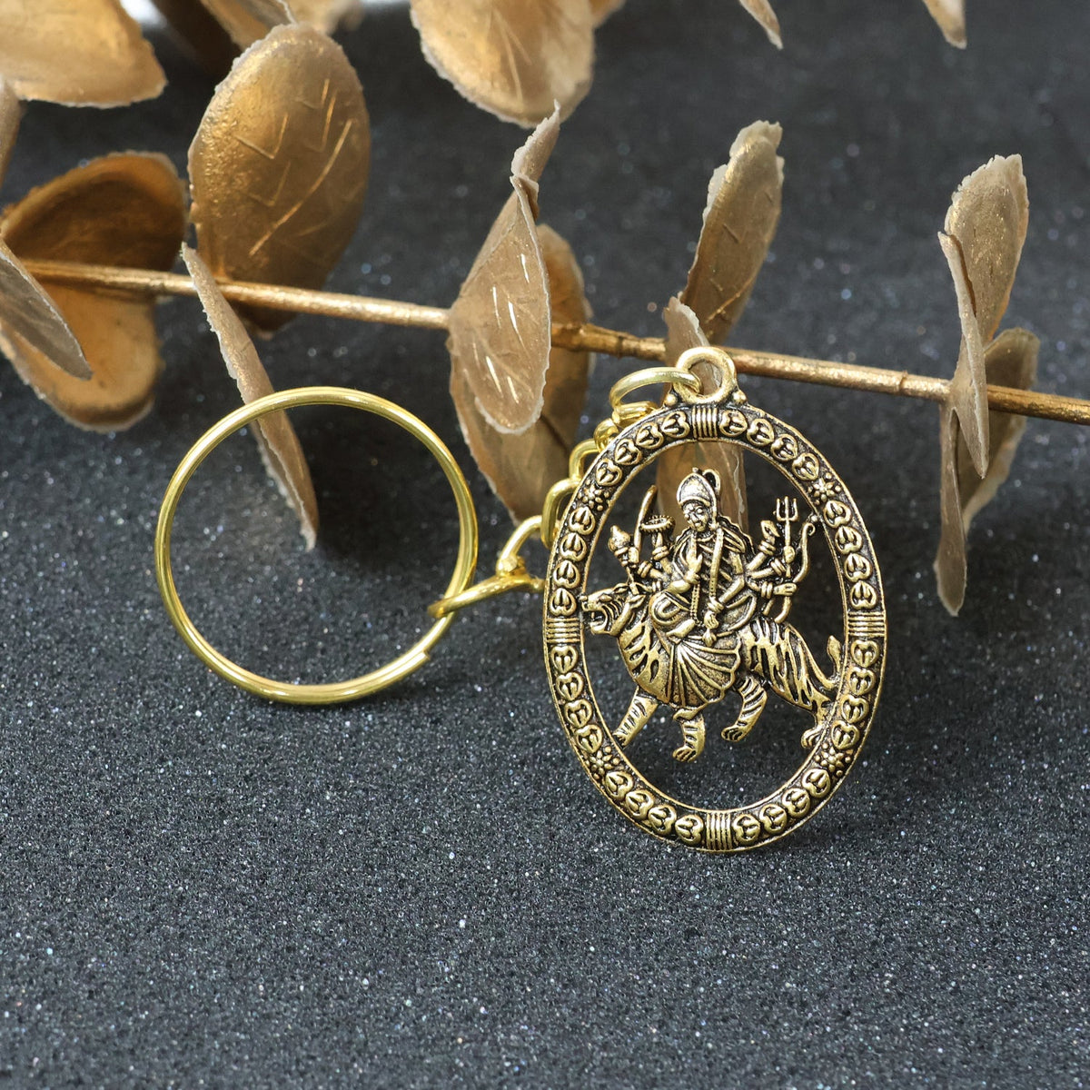 A golden keychain with an oval pendant showing a deity, placed on a dark textured surface with golden dried leaves.
