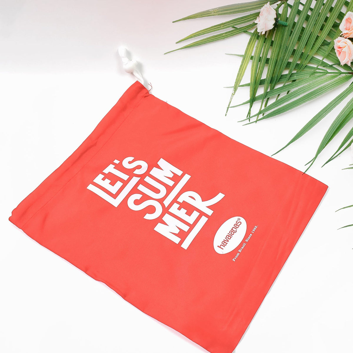 A red drawstring bag with "LET'S SUMMER" and Havaianas logo, angled on a white surface with palm leaves and small pink