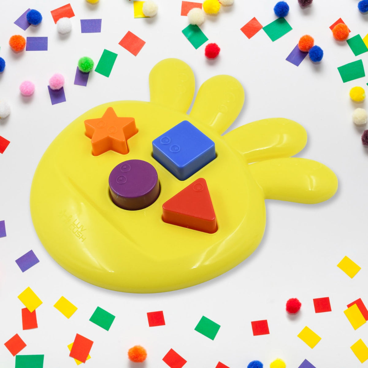 Yellow shape sorter with four colorful blocks on a white surface, surrounded by scattered confetti and pom-poms.