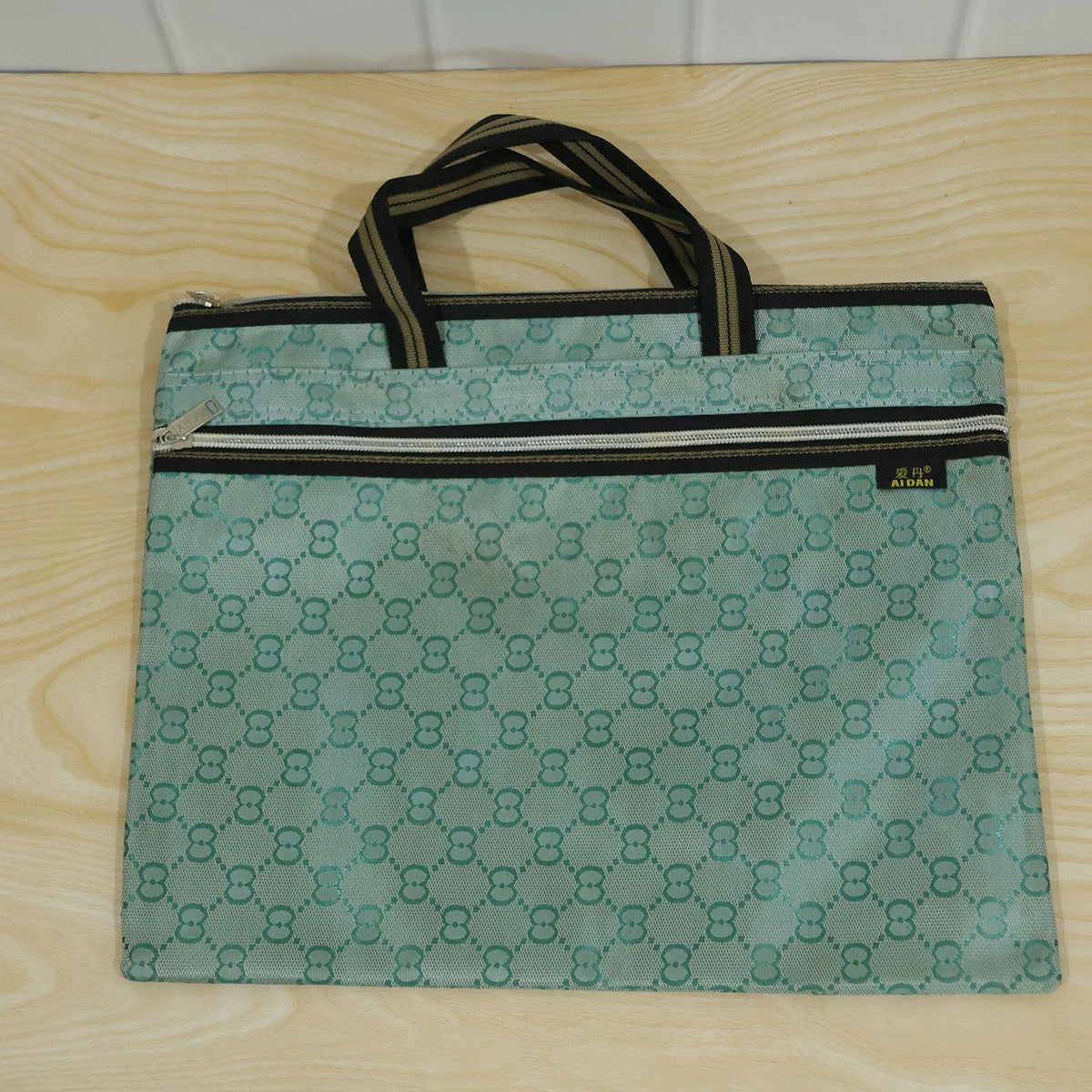 A green patterned document bag with black and gold striped handles and two zippers, resting flat on a light wooden surface.