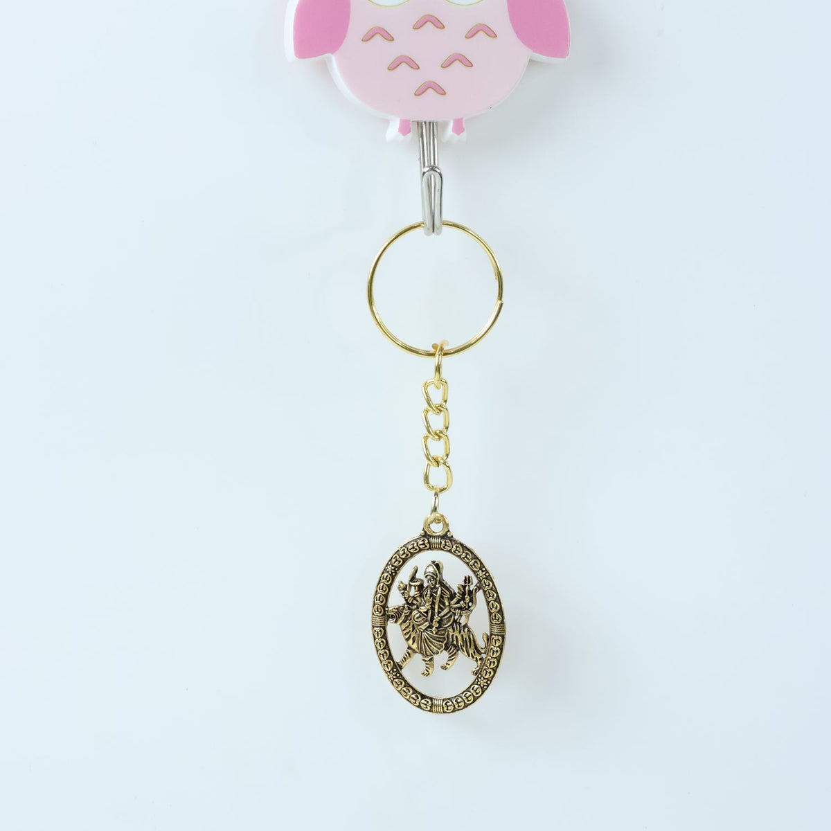 A golden keychain with an oval deity pendant hangs from a pink owl-shaped hook on a plain white wall.