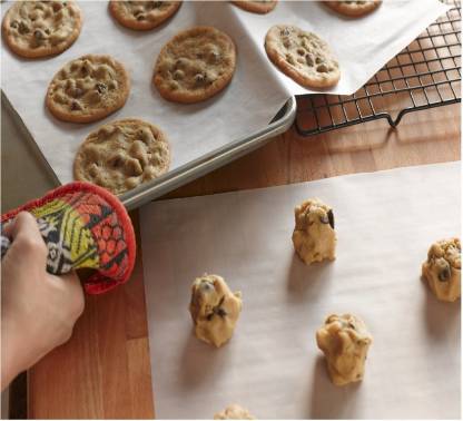 A hand in a colorful oven mitt next to cookie dough balls on baking paper and baked cookies on a tray.