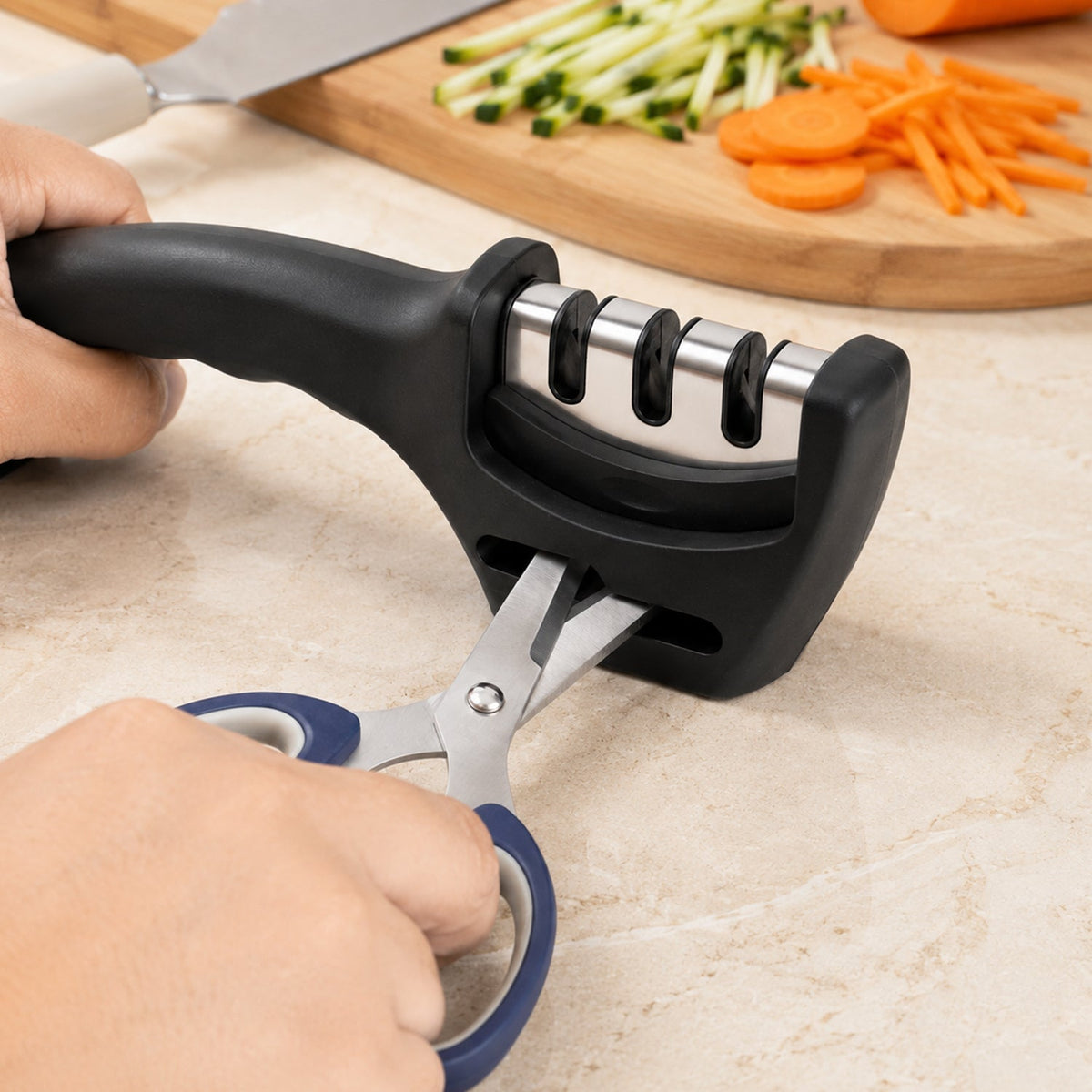 Hands hold a black three-stage sharpener, sharpening blue-handled scissors with chopped vegetables in the background.