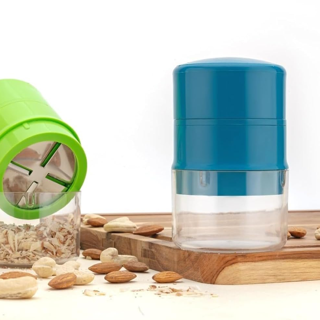A green dry fruit slicer with sliced nuts in its base lies on a wooden board next to a blue and clear slicer.