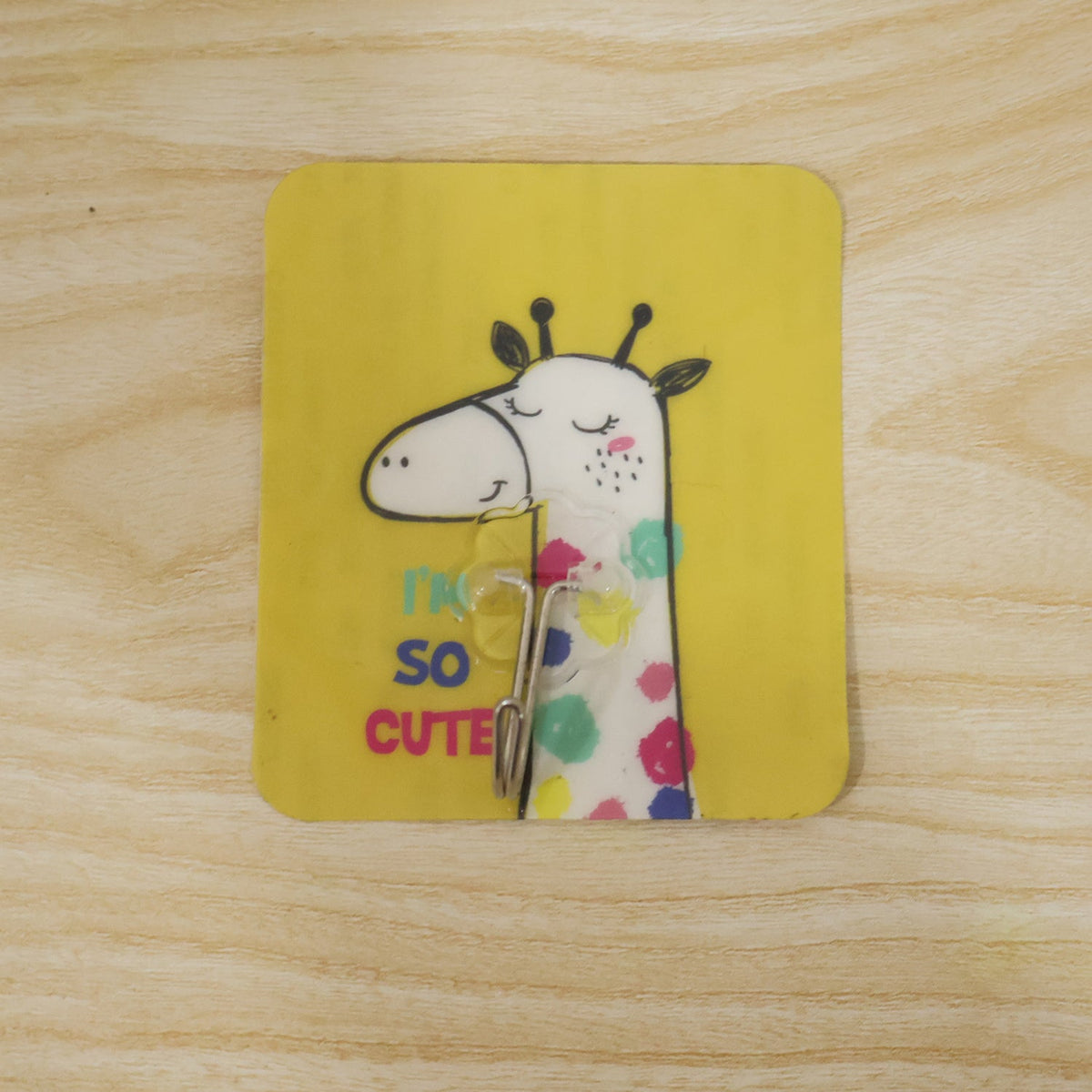 A yellow square wall hook with a cartoon giraffe and text "I'M SO CUTE" is adhered to a light wooden surface.