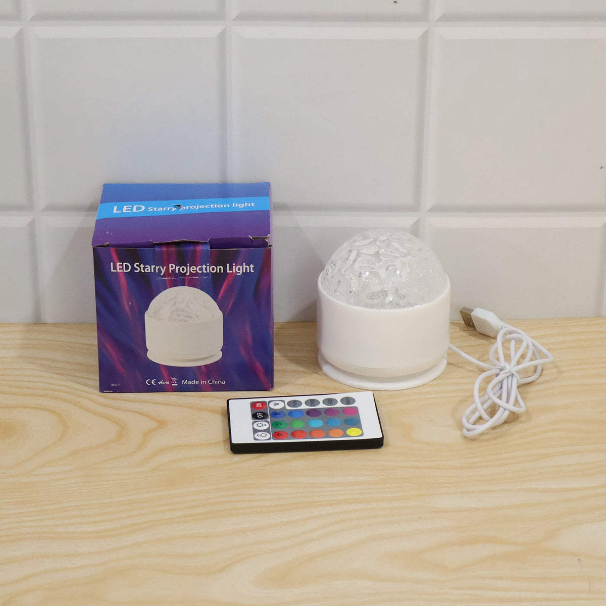 A white LED projection light, its box, and a remote control on a light wooden surface against a white tiled wall.