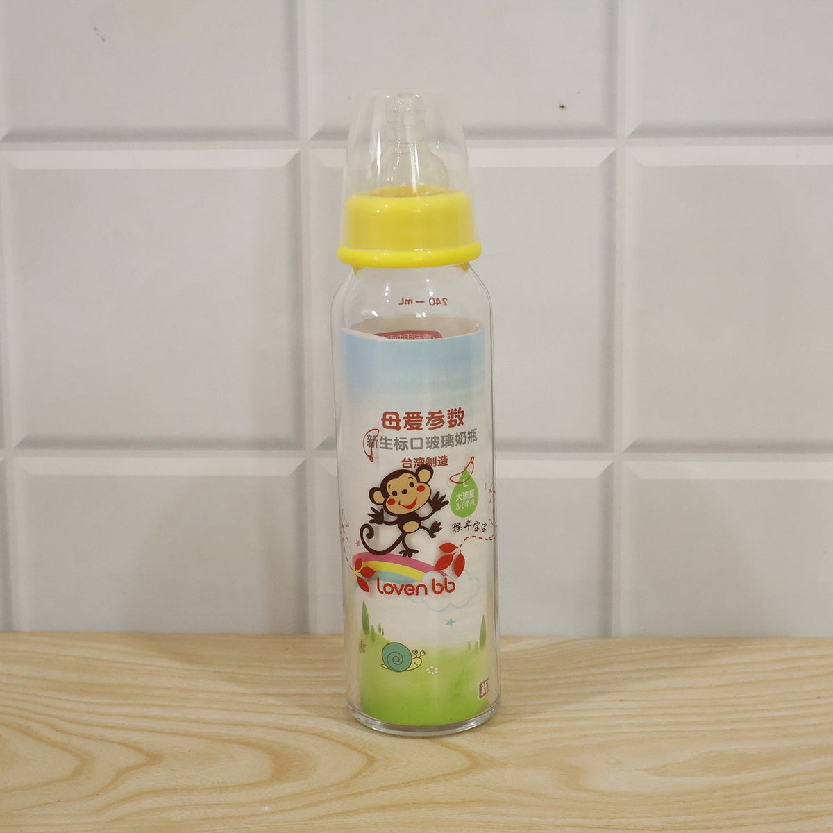 A clear glass baby bottle with a yellow nipple and a colorful monkey and snail design stands on a wooden surface.