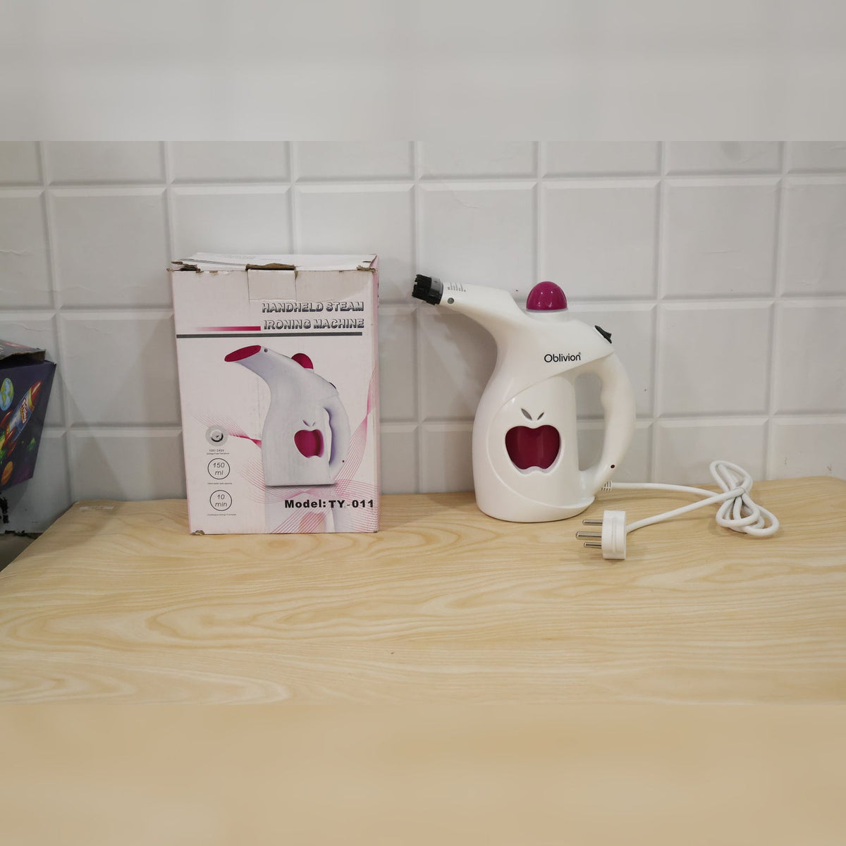A white and pink handheld steamer with its packaging box, both featuring an apple logo, on a wooden surface.