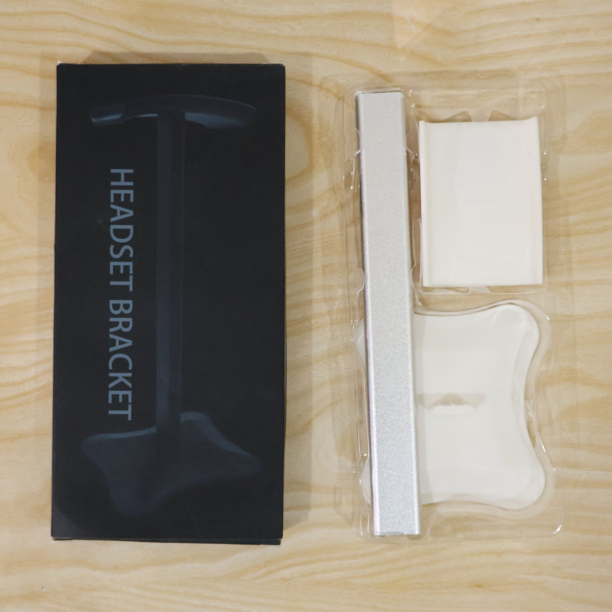 Disassembled silver and white headphone stand components in clear plastic packaging next to its black box.