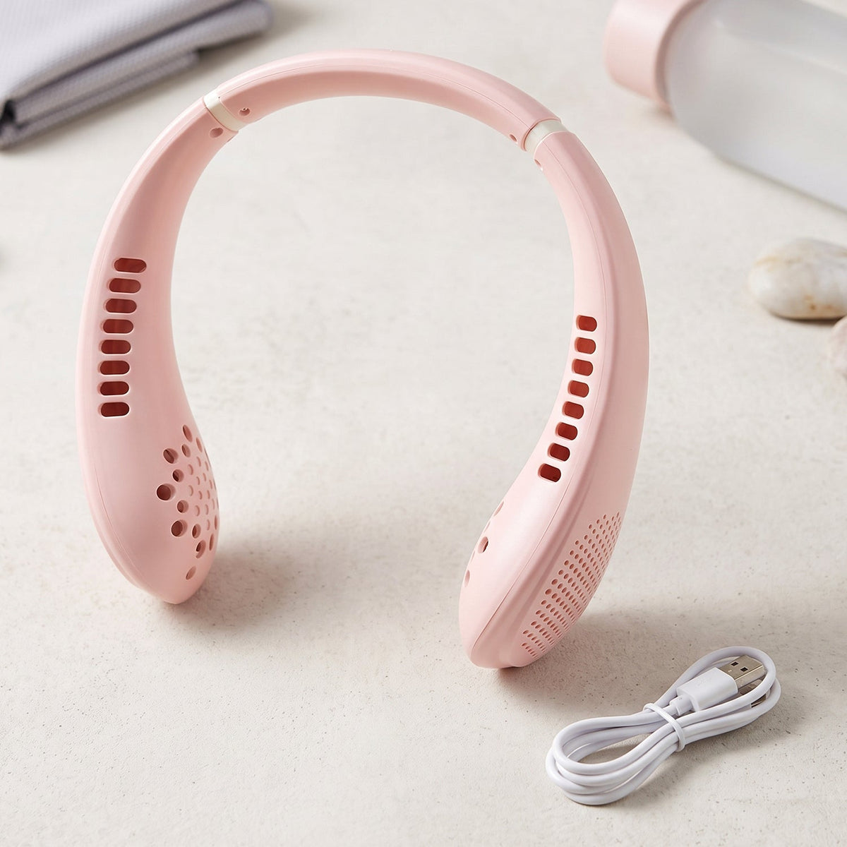 A pink portable neck fan with a white USB charging cable on a light surface with blurred background items.