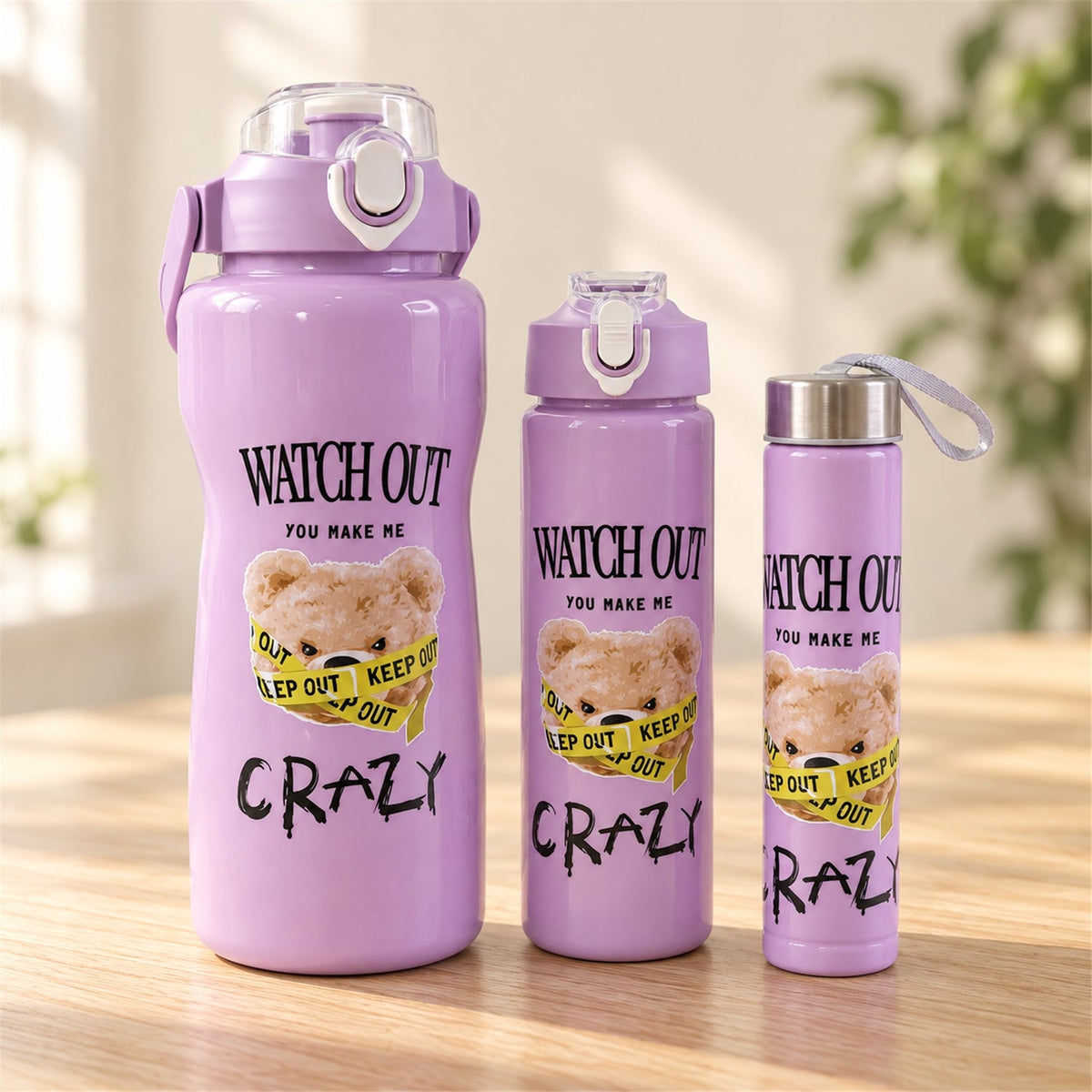 Three purple water bottles with a teddy bear design, in varying sizes, on a wooden table with a blurred green plant in the