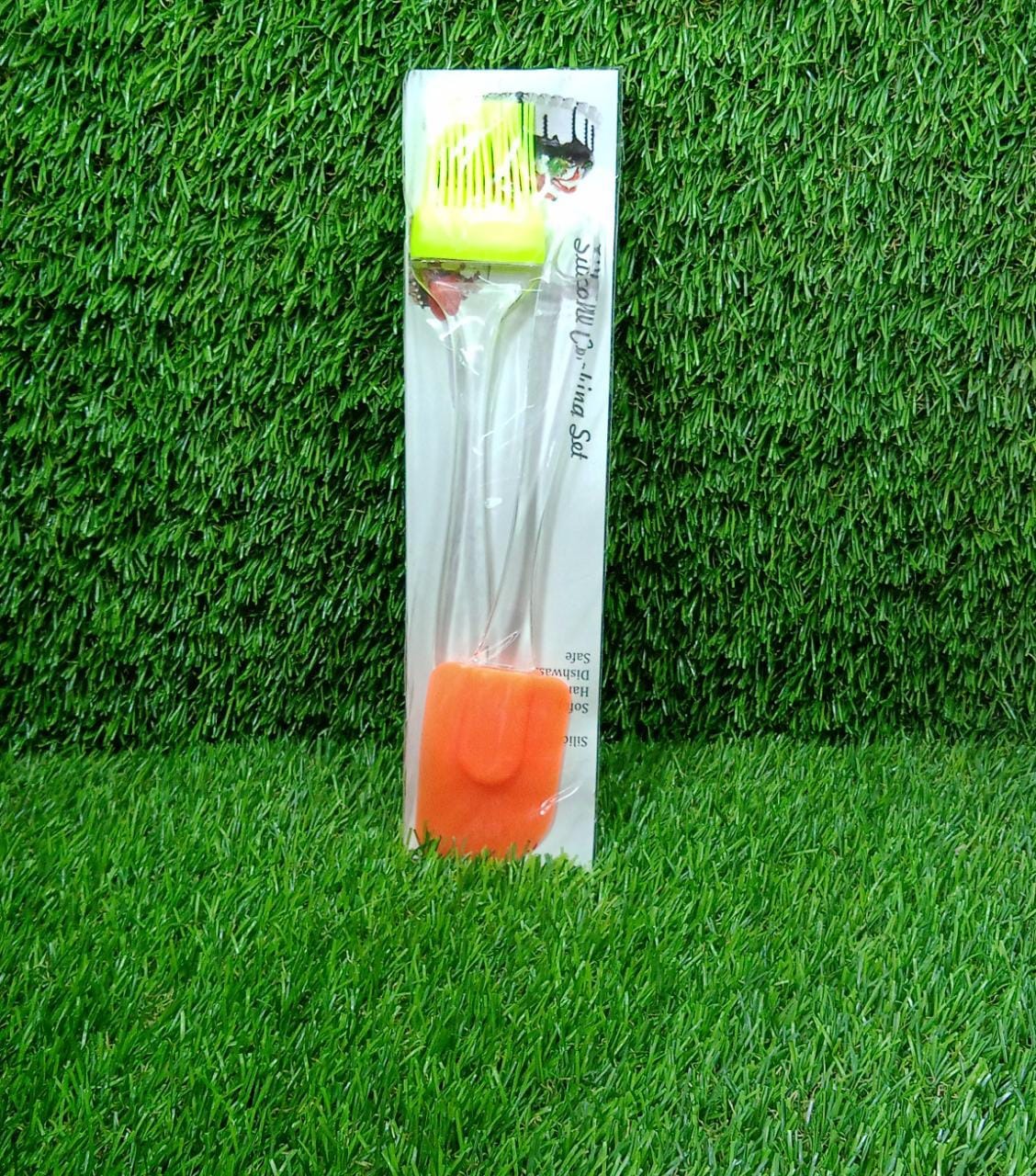 A packaged set containing an orange silicone spatula and a green silicone brush stands upright on a vibrant green grassy