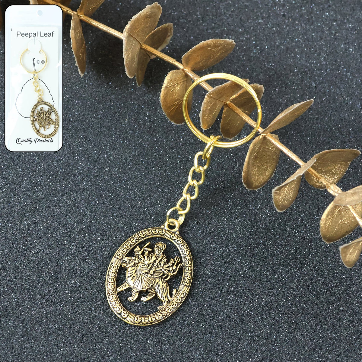 A golden keychain featuring an oval pendant with a deity design, positioned on a dark surface beside dried golden foliage.