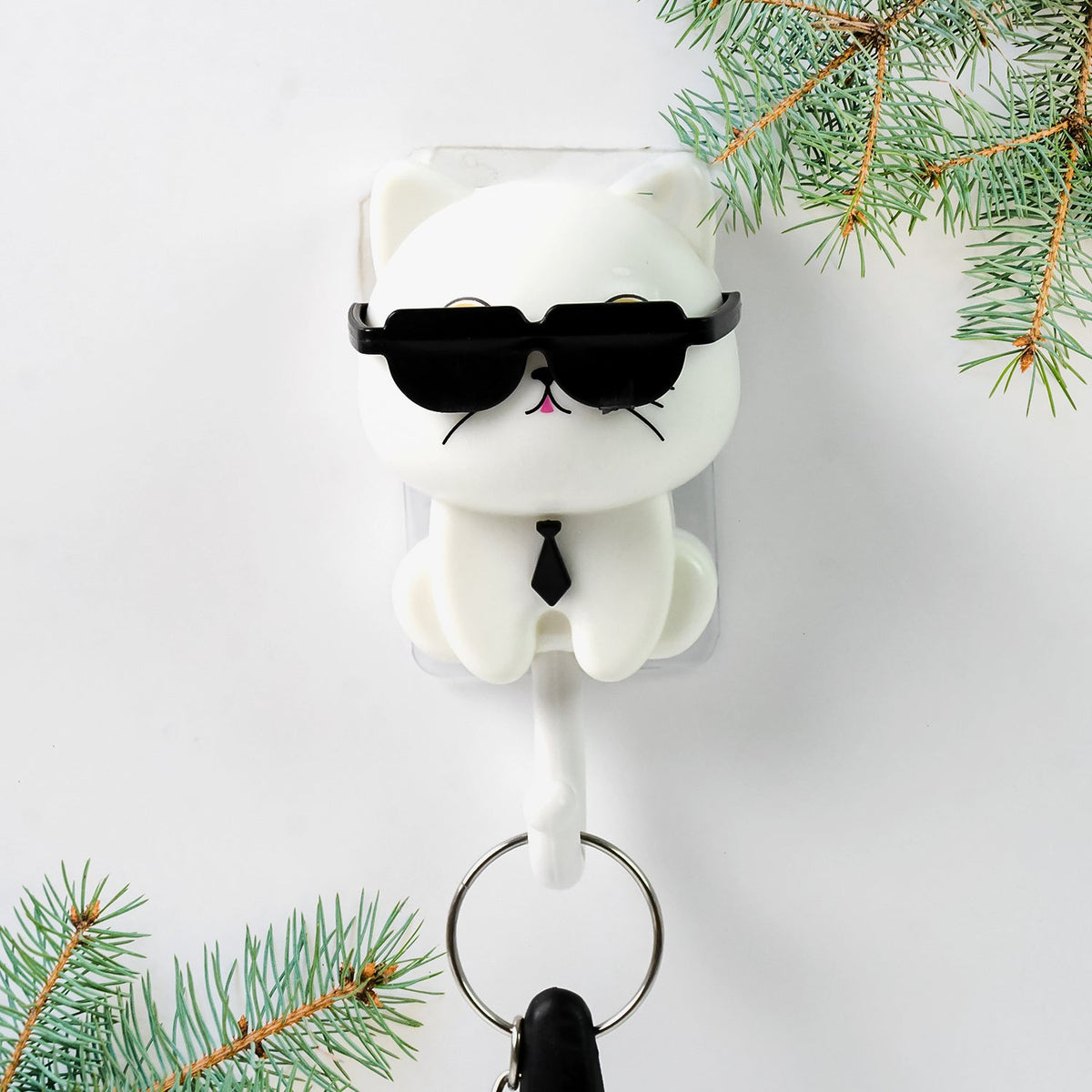 White cat-shaped wall hook with sunglasses and a tie, holding a key ring, surrounded by green pine branches.
