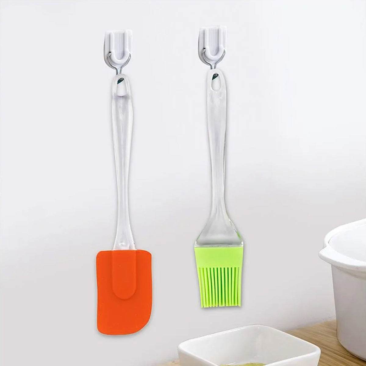 An orange silicone spatula and a green silicone basting brush hang side-by-side on white adhesive hooks against a white wall.