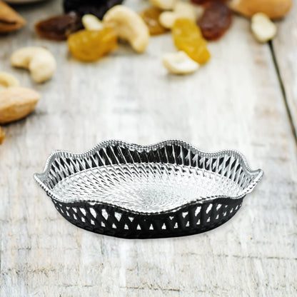 A silver plastic scalloped serving bowl with a textured bottom sits on a light wooden surface, surrounded by various nuts