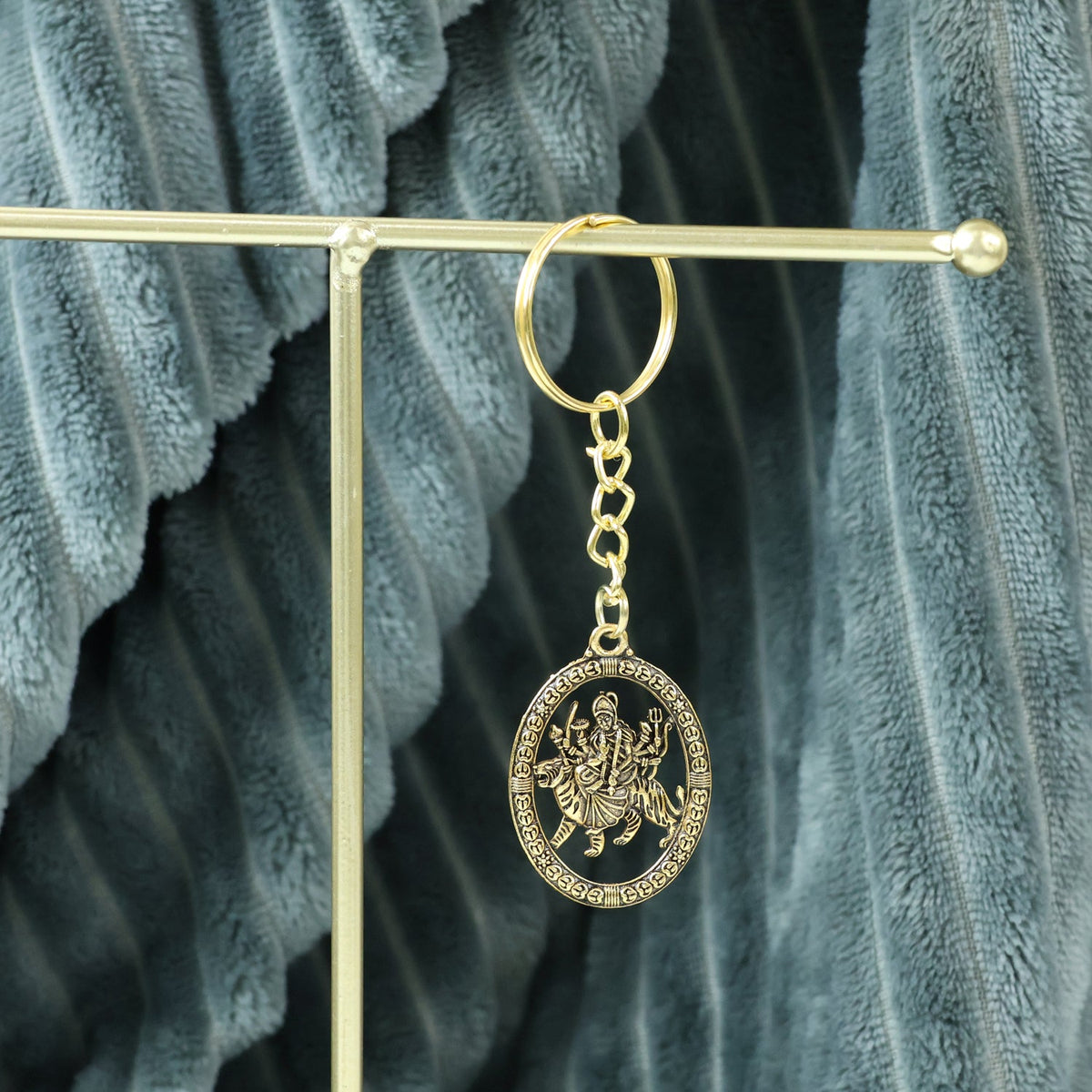 A golden keychain with an oval deity pendant hangs from a thin golden rod against a soft gray textured fabric.
