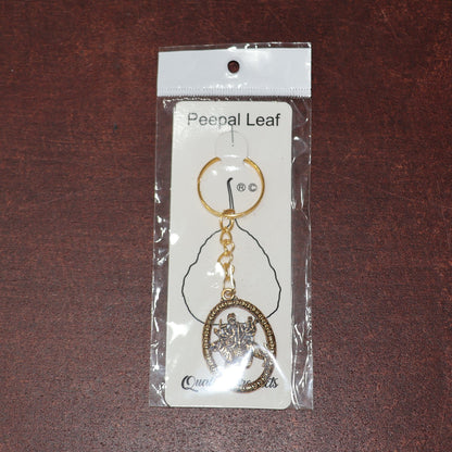 A golden keychain with an oval deity pendant, sealed within clear retail packaging on a brown leather surface.