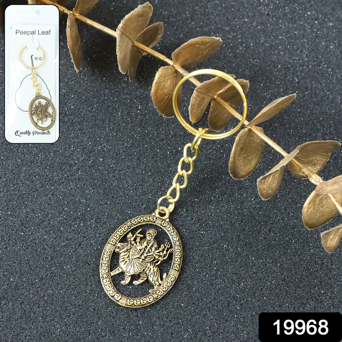 A golden keychain with an oval deity pendant, displayed in clear packaging against a dark background with dried leaves.