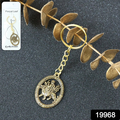 A golden keychain with an oval deity pendant, displayed in clear packaging against a dark background with dried leaves.