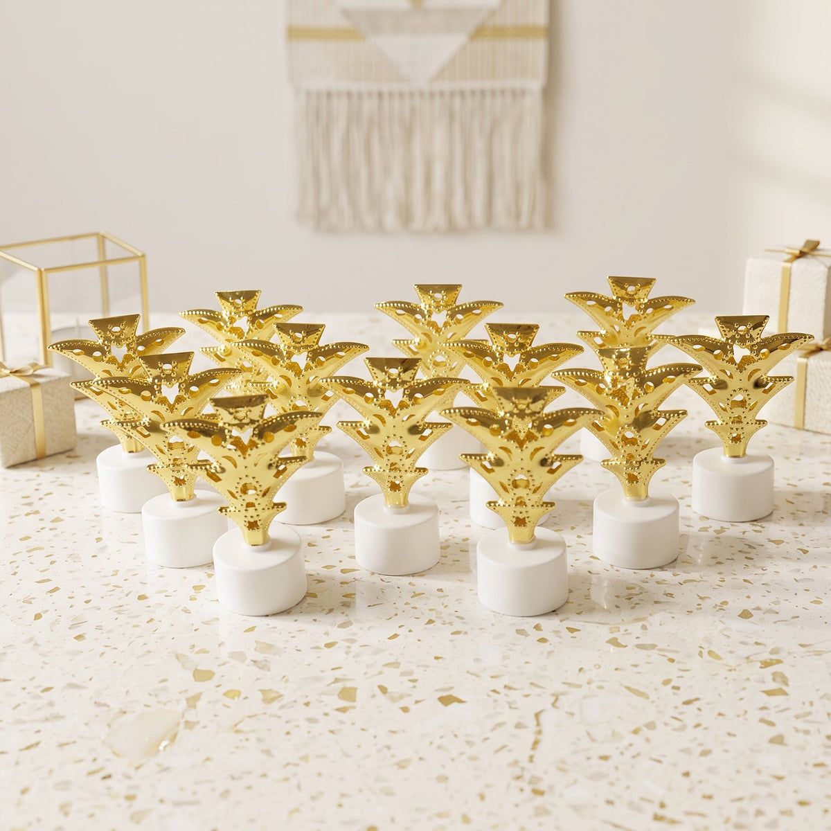 A dozen golden Christmas tree LED tealights stand on a white speckled surface, surrounded by gift boxes.