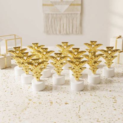 A dozen golden Christmas tree LED tealights stand on a white speckled surface, surrounded by gift boxes.