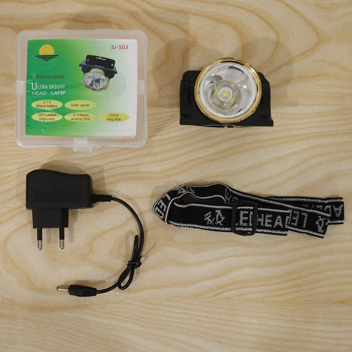 A black and gold headlamp, a charger, a head strap, and a clear box with product information on a light wooden surface.
