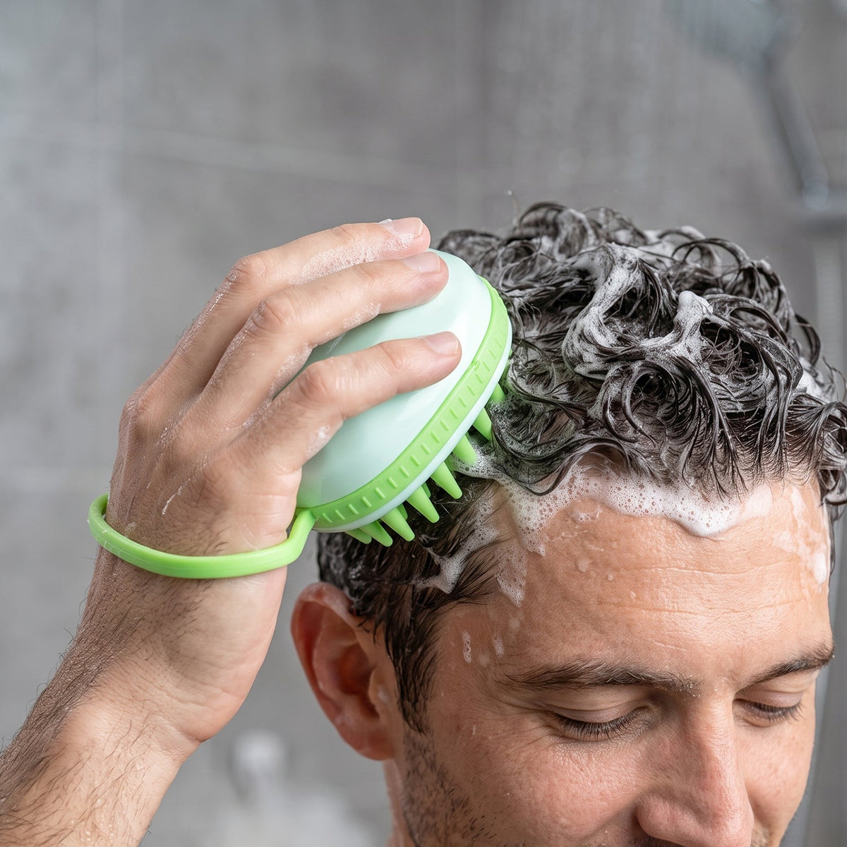 A person's hand is visible, holding a light green silicone scalp massage brush and scrubbing their dark, sudsy hair in a