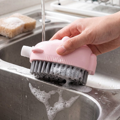 A hand holds a pink bear-shaped soap dispensing brush, scrubbing a stainless steel sink under running water.