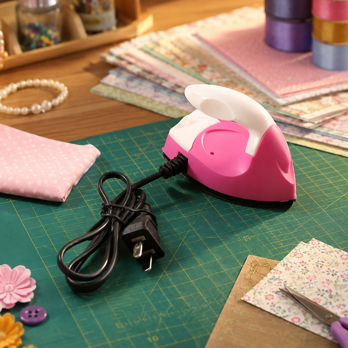A pink and white mini iron with its black cord and plug sits on a green cutting mat surrounded by craft supplies.