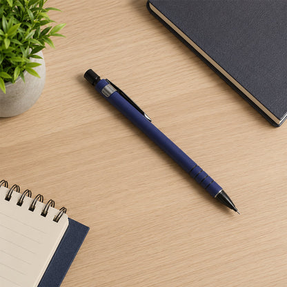 A blue mechanical pencil lies on a light wooden desk next to a green plant, a dark blue notebook, and a spiral notepad.