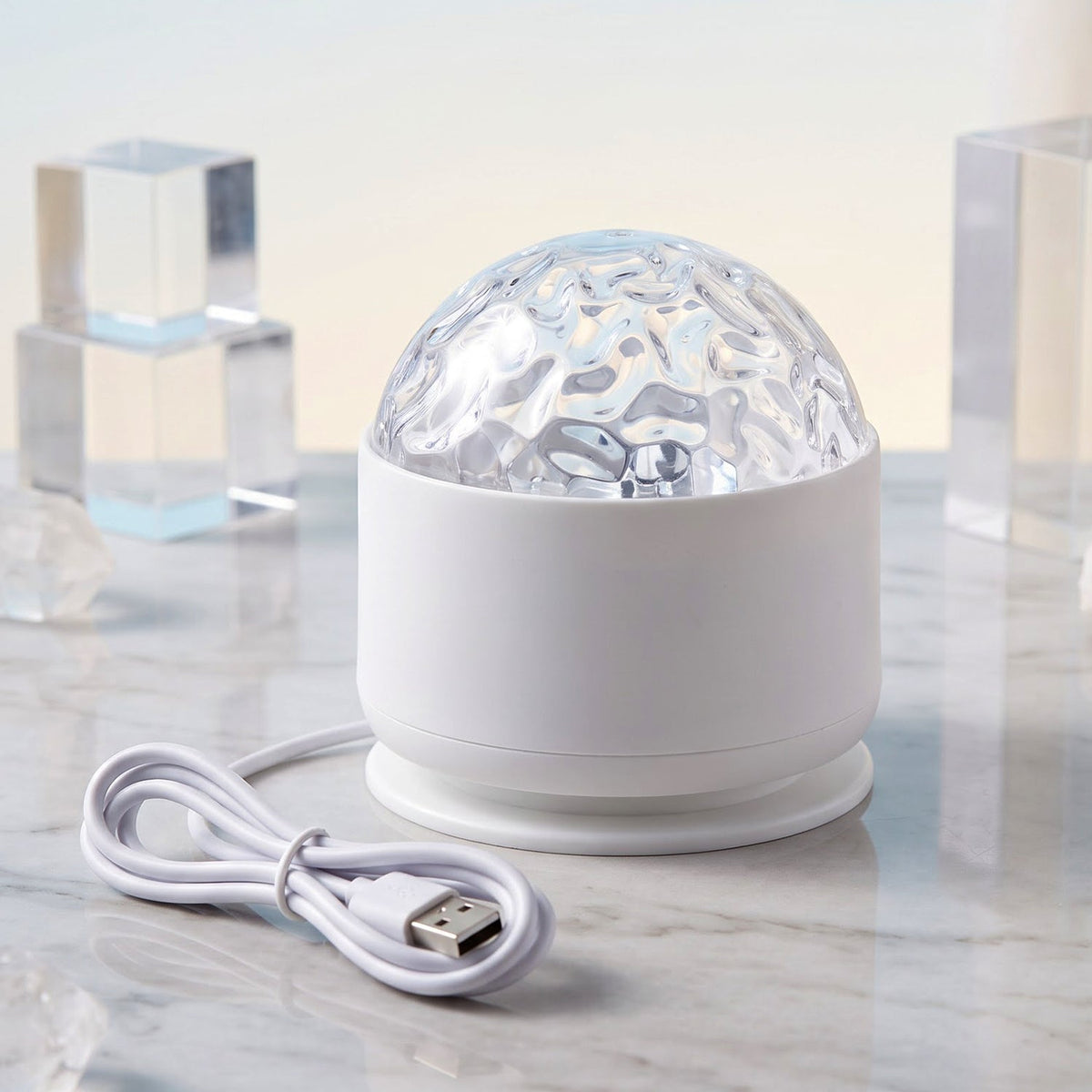 A white LED projection light with a clear textured dome and a white USB cable on a marble surface with clear blocks.