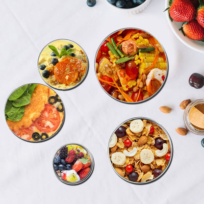 Four open stainless steel containers filled with diverse meals, including fruit, cereal, and a chicken dish, arranged on a