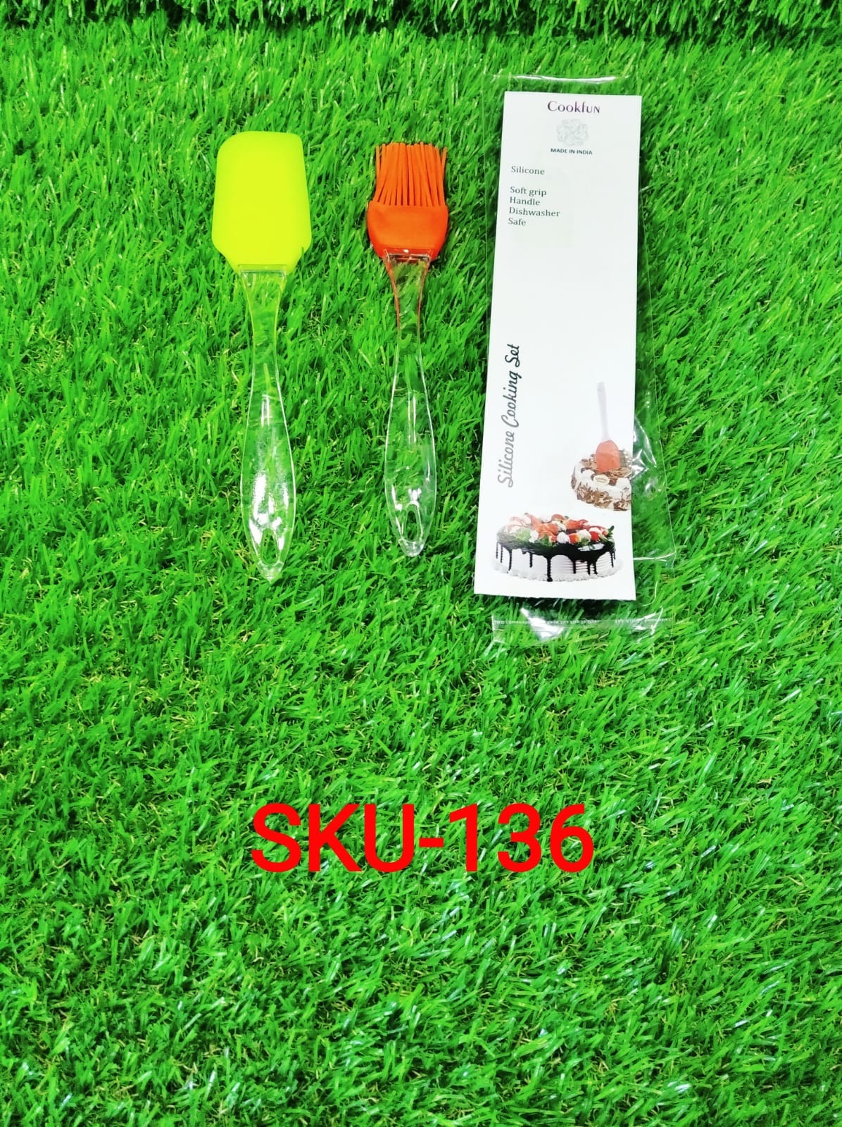 A yellow silicone spatula, an orange silicone brush, and a packaged silicone cooking set lie on artificial green grass.