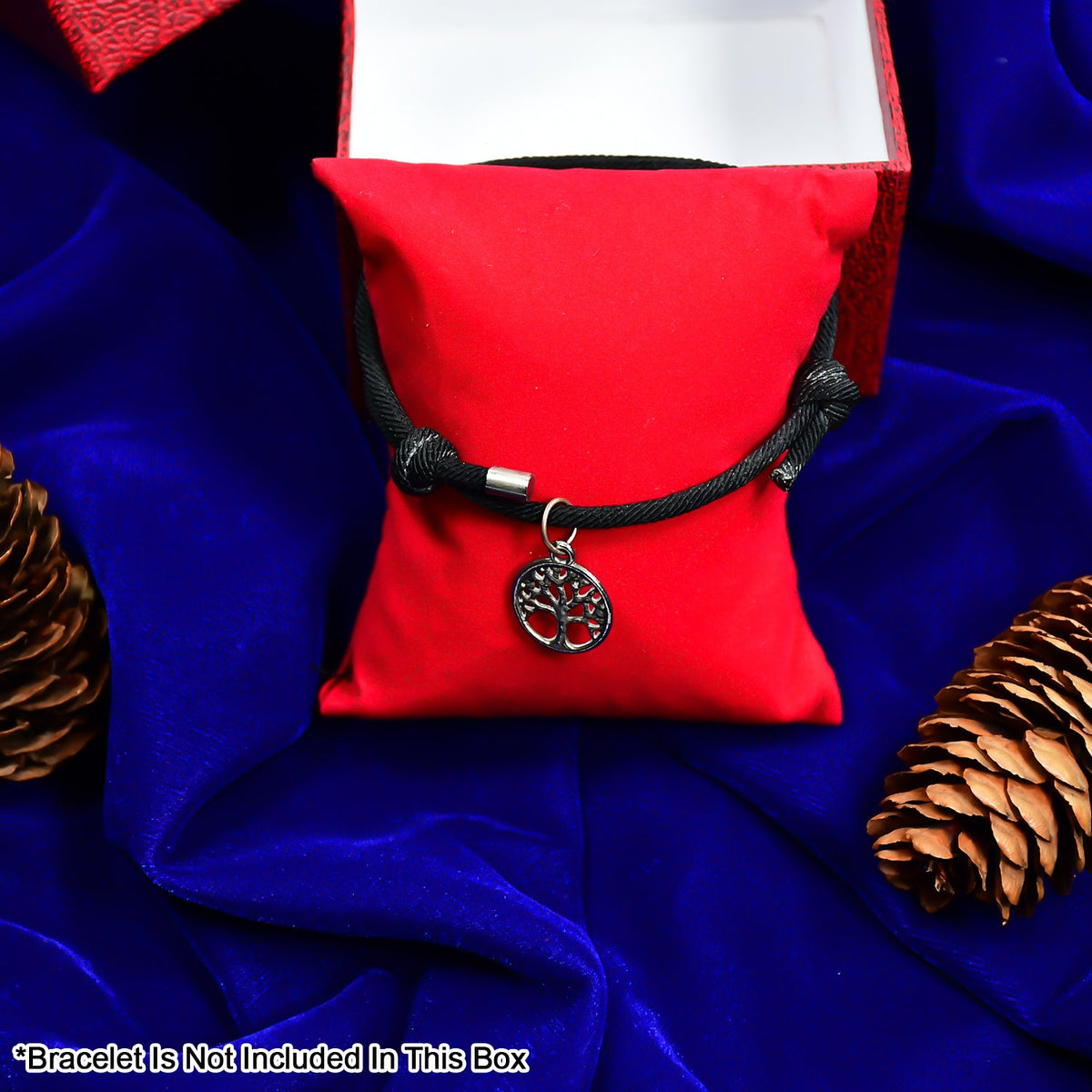 A black cord bracelet with a silver tree charm displayed on a red velvet pillow, surrounded by pinecones on a blue cloth.