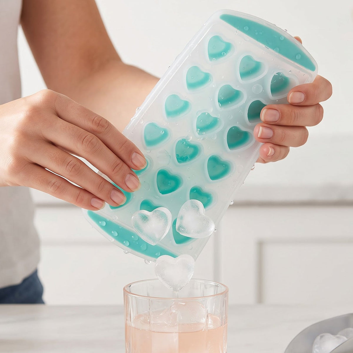 A person's hands gently twist a clear and teal heart-shaped ice cube tray, releasing two heart-shaped ice cubes – view 3