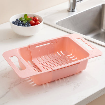 Pink plastic adjustable over-sink drain basket on a white countertop with a bowl of berries and a sink in the background.