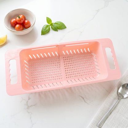 Pink plastic adjustable over-sink drain basket on a white marble countertop with a bowl of tomatoes and basil.