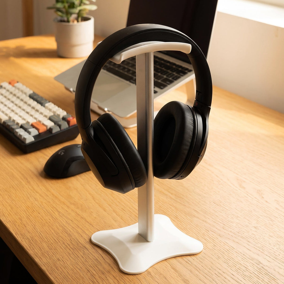 White and silver headphone stand on a light wooden desk, with a grey laptop and mechanical keyboard nearby.
