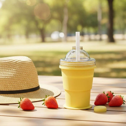 A yellow silicone cup with a straw and dome lid sits on a wooden table outdoors with strawberries and a straw hat.