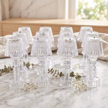 Twelve clear acrylic miniature lamps with ribbed shades are arranged on a white marble surface with delicate white flowers.