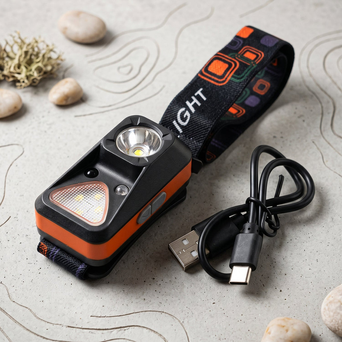 The orange and black headlamp with its strap and a coiled USB-C charging cable, surrounded by small rocks and a compass on a