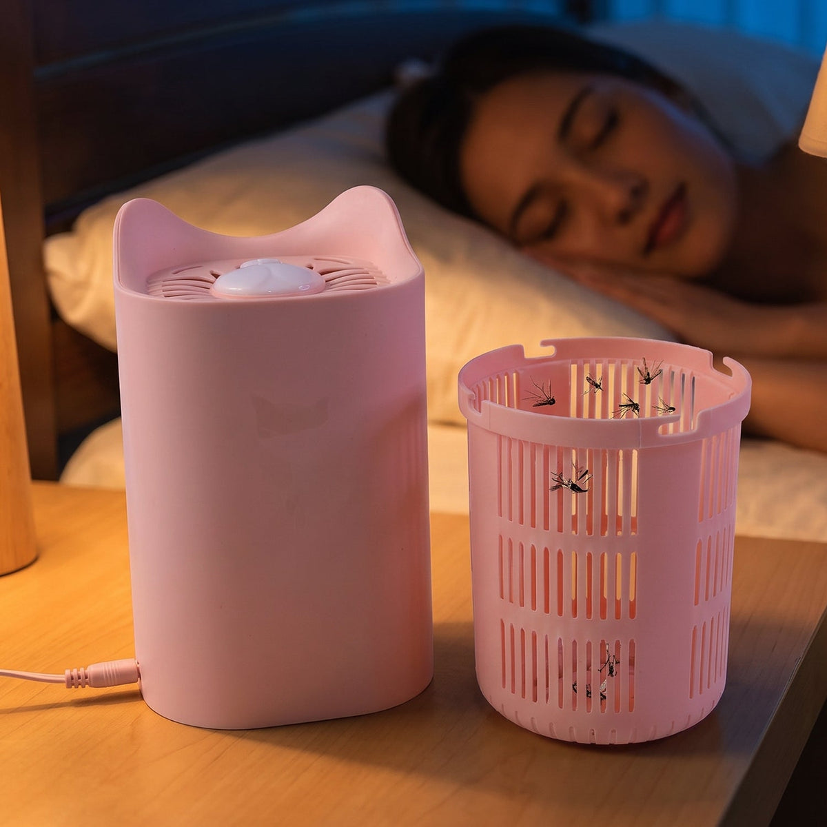 A pink cat-shaped mosquito lamp and its collection basket with trapped mosquitoes sit on a nightstand while a woman sleeps.