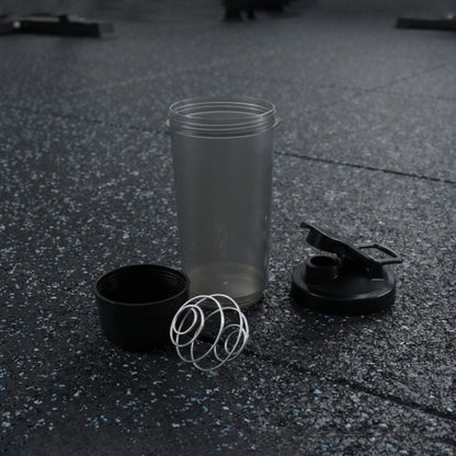 A disassembled grey and black shaker bottle with a metal whisk is on a dark gym floor.