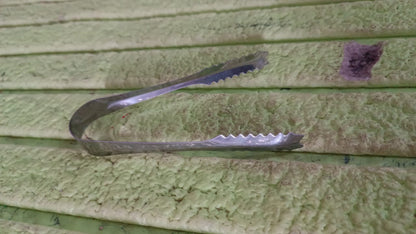 A silver tong rests on a textured light green surface with horizontal ridges.