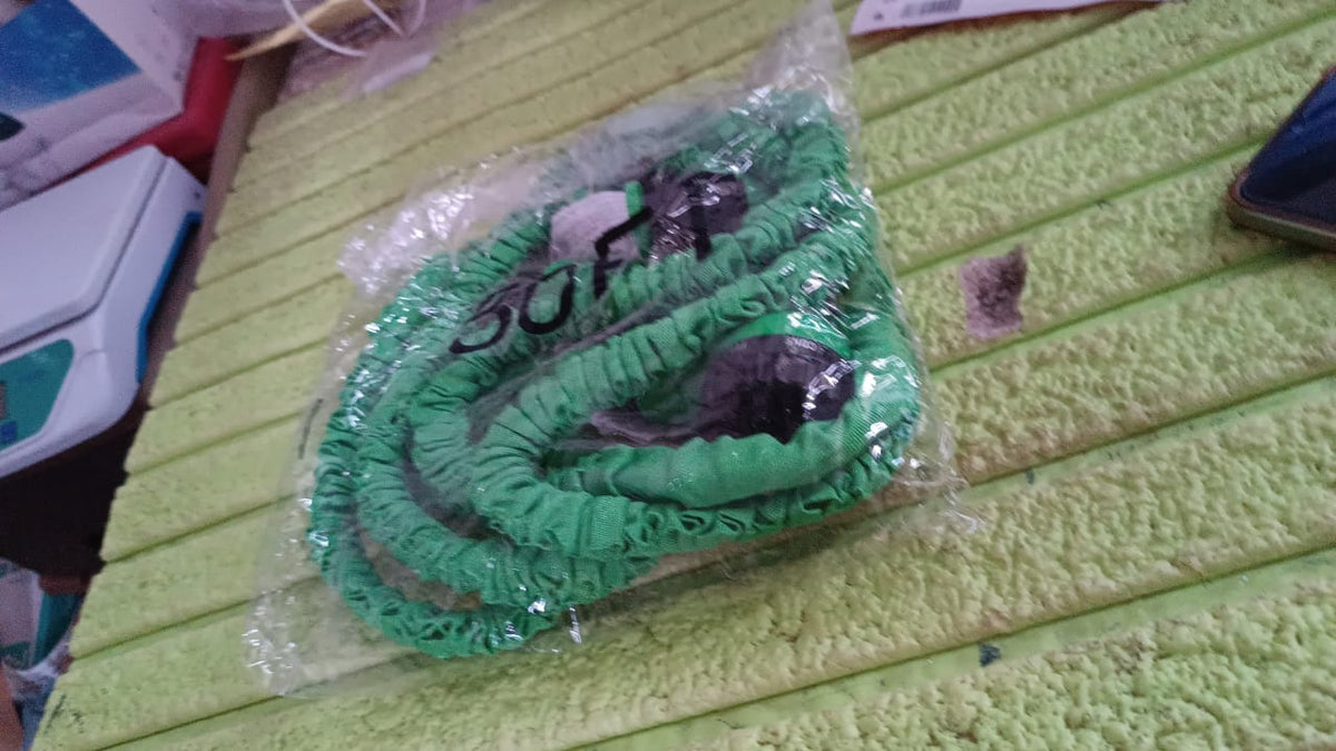 A green expandable garden hose, coiled and sealed in clear plastic packaging, resting on a textured green surface.